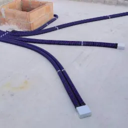 Niebieskie rury rekuperacji prowadzone po jasnoszarym dachu w kierunku komina z cegły, widoczne łączenia i zakończenia rur.