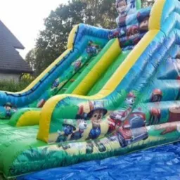 Duży, kolorowy dmuchany zamek ze zjeżdżalniami z motywem bajki Psi Patrol, rozłożony na trawie i zabezpieczony niebieską plandeką.