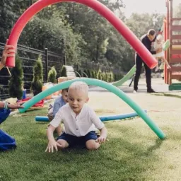 Kobieta w białej koszulce i niebieskiej spódnicy bawi się z dziećmi na trawie, używając kolorowych piankowych rurek do stworzenia tunelu.