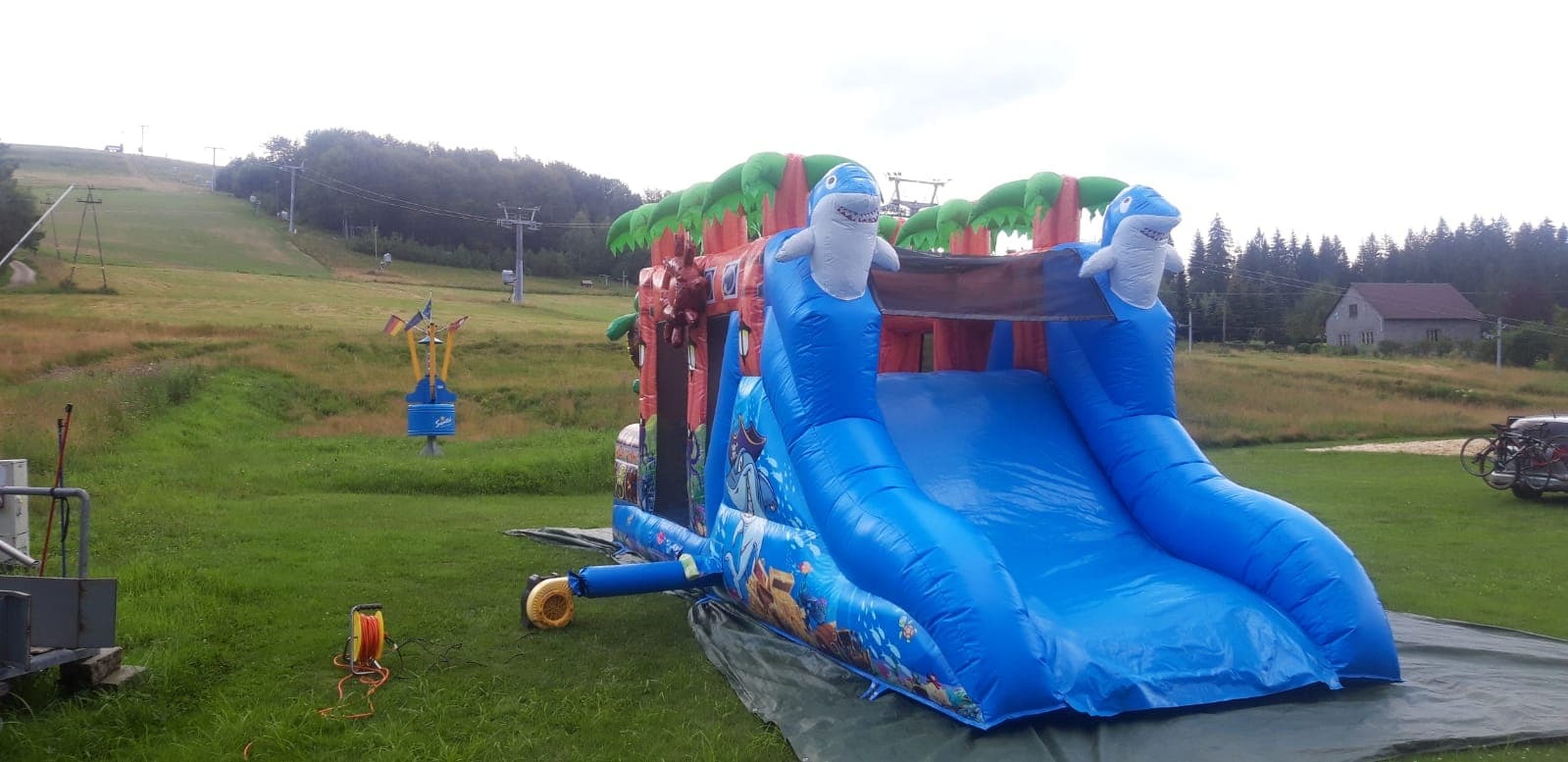 Niebieska dmuchana zjeżdżalnia z motywem morskim i rekinami, ustawiona na zielonym trawniku w pobliżu stoku narciarskiego i domów.