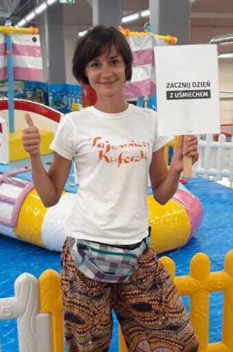 Uśmiechnięta kobieta w koszulce z napisem 'Tajemniczy Kuferek' trzymająca tabliczkę z napisem 'Zacznij dzień z uśmiechem', w tle kolorowa konstrukcja przypominająca statek piracki na placu zabaw.