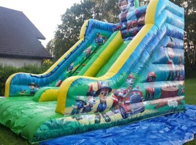 Duży, kolorowy dmuchany zamek ze zjeżdżalniami z motywem bajki Psi Patrol, rozłożony na trawie i zabezpieczony niebieską plandeką.