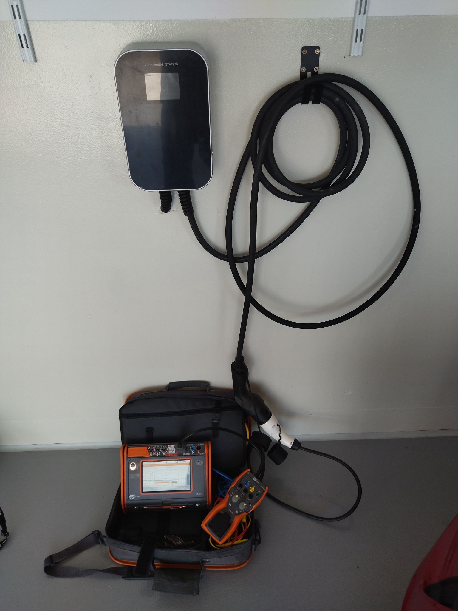 Zamontowana na ścianie stacja ładowania pojazdów elektrycznych z podłączonym kablem, obok leży otwarty futerał z miernikiem instalacji elektrycznej.