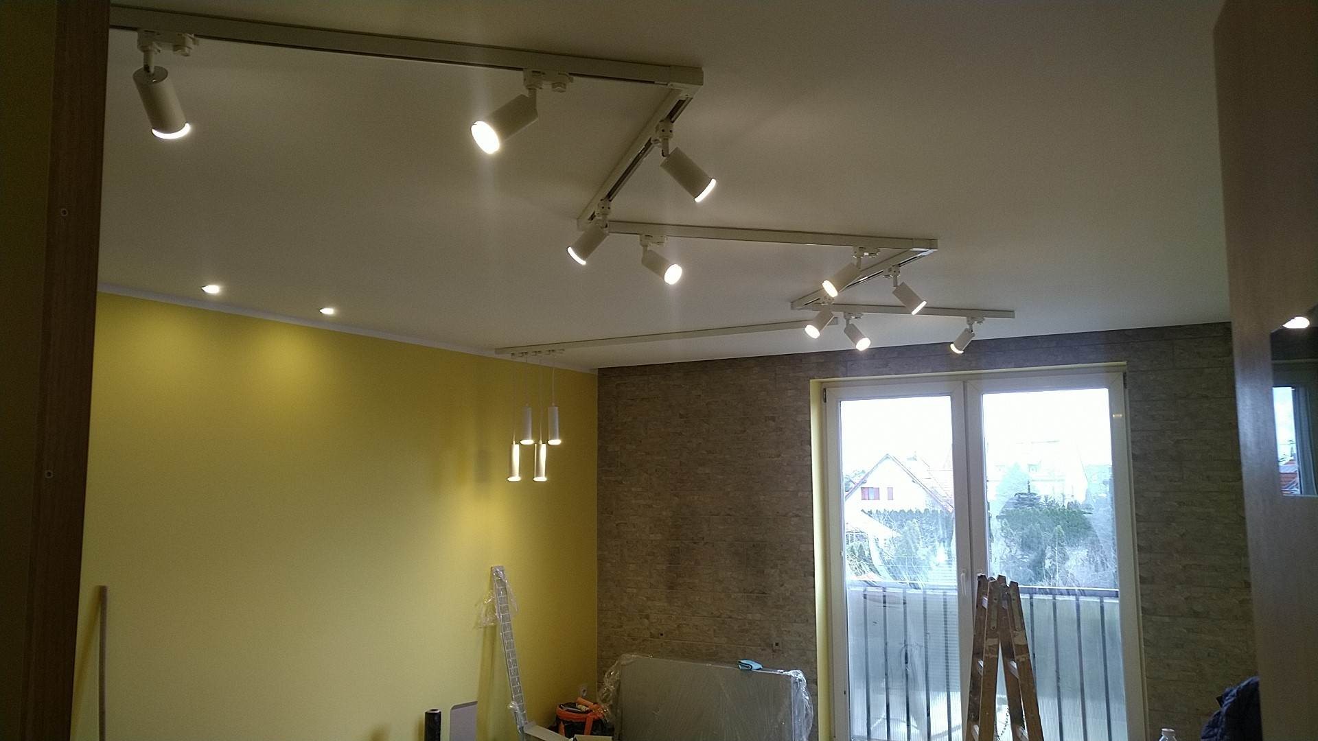 Wnętrze pokoju z zamontowanym systemem szynowym oświetlenia punktowego, skierowane na żółtą ścianę i okno balkonowe z widokiem na zewnątrz. Dodatkowo widoczne wiszące lampy cylindryczne.