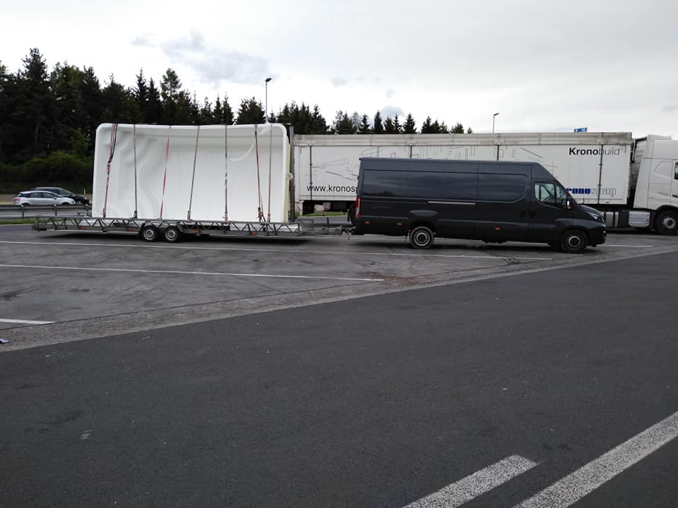 Czarny van Iveco ciągnie przyczepę z zabezpieczonym białym ładunkiem, prawdopodobnie kontenerem lub modułową konstrukcją, na parkingu z ciężarówkami w tle.