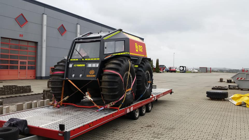 Amfibia Sherp w kolorze czarnym z żółtymi akcentami i numerem 112, zabezpieczona pasami transportowymi na niskopodwoziowej przyczepie z czerwonymi elementami, na tle szarego budynku z czerwonymi...