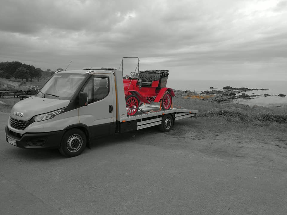 Grey Iveco laweta przewozi jaskrawoczerwony, zabytkowy samochód osobowy na tle pochmurnego wybrzeża.