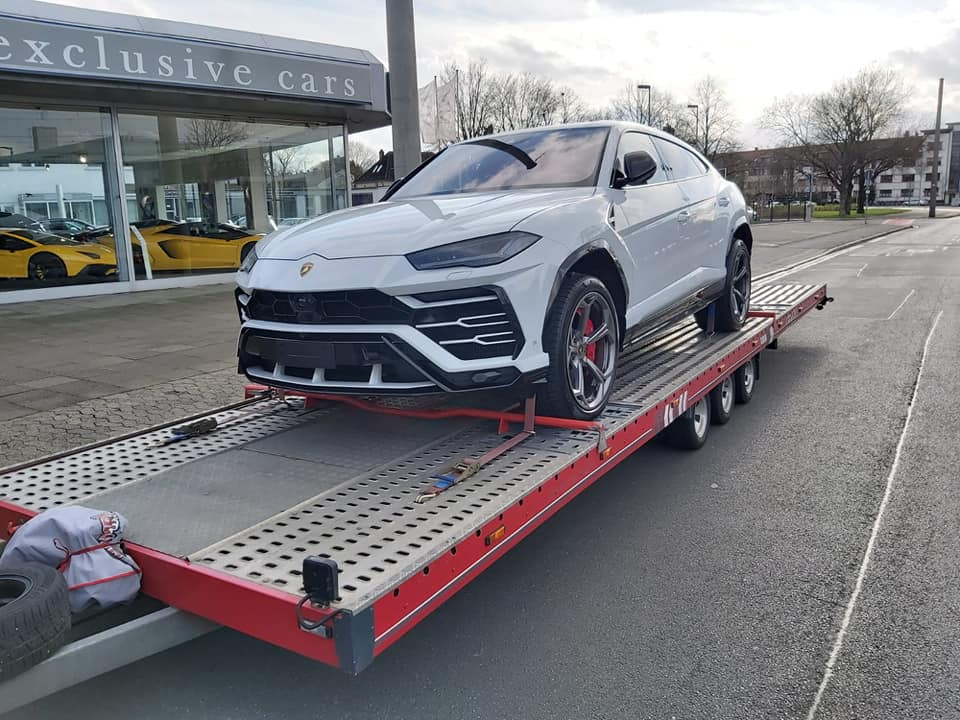 Biały Lamborghini Urus zabezpieczony pasami na czerwonej lawecie, w tle budynek salonu samochodowego Exclusive Cars z widocznymi żółtymi sportowymi autami.