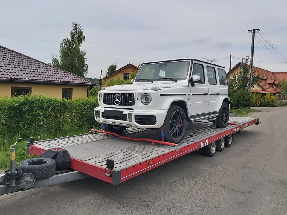 Biały Mercedes-Benz G-klasy zabezpieczony pasami na czerwonej lawecie z perforowaną platformą, w tle domy jednorodzinne i słup elektryczny.