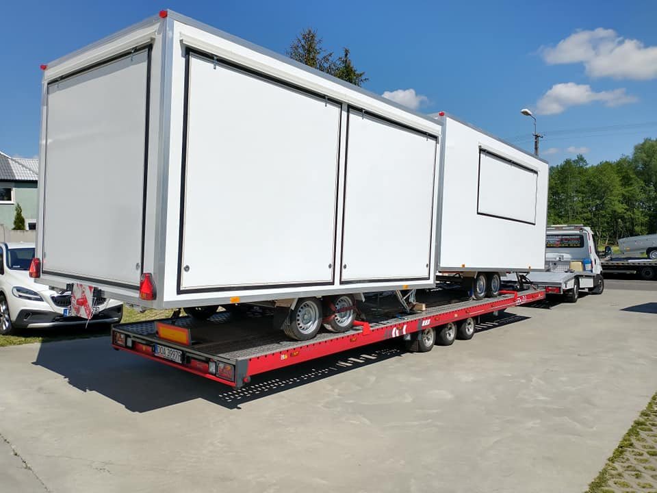 Laweta z platformą w kolorze czerwonym transportuje biały kontener z przesuwnymi drzwiami w słoneczny dzień.