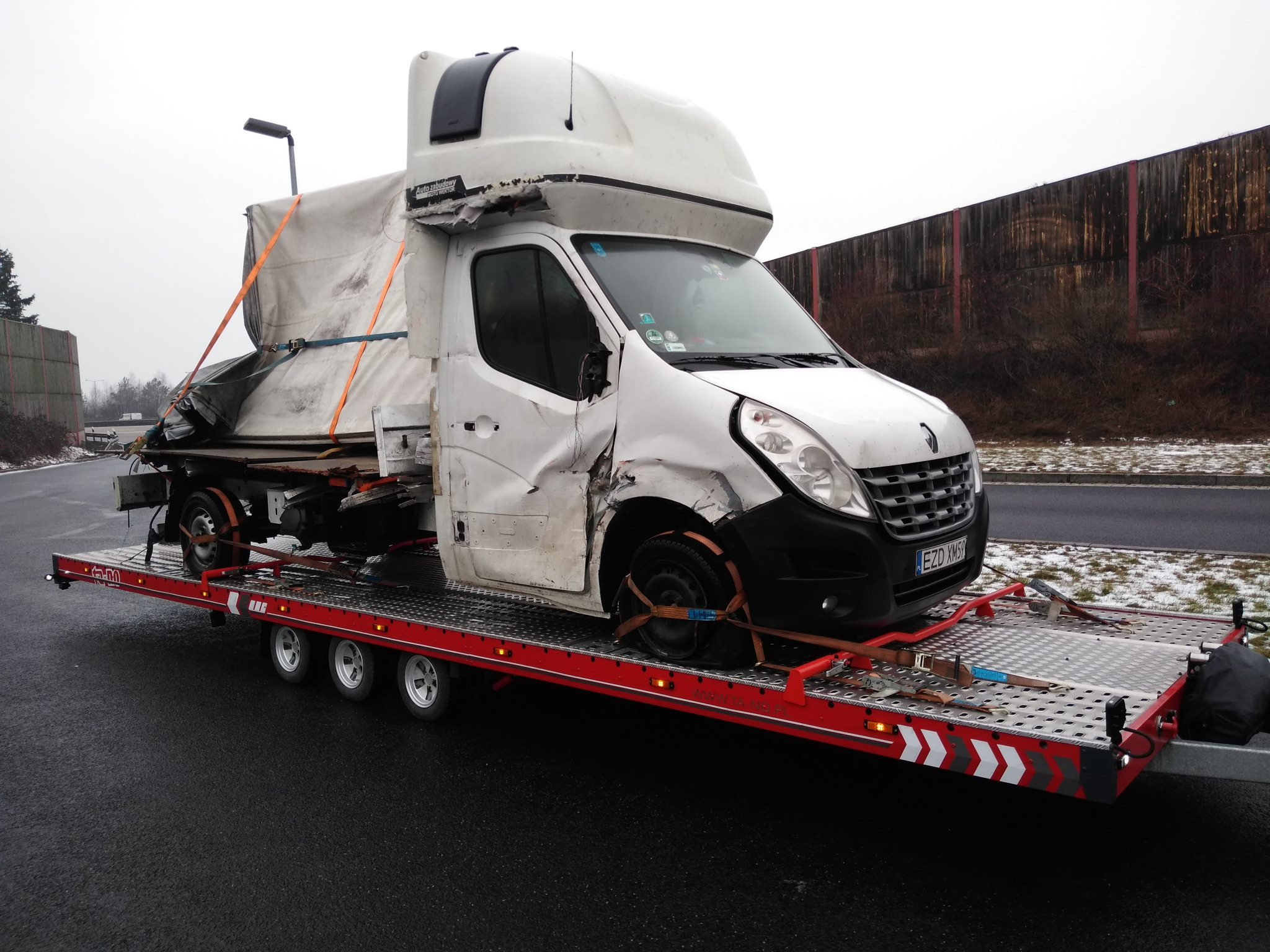 Uszkodzony biały samochód dostawczy Renault z plandeką, z widocznymi wgnieceniami i uszkodzeniami karoserii, zabezpieczony pasami transportowymi na czerwonej lawecie z trzema osiami, na tle szarej...