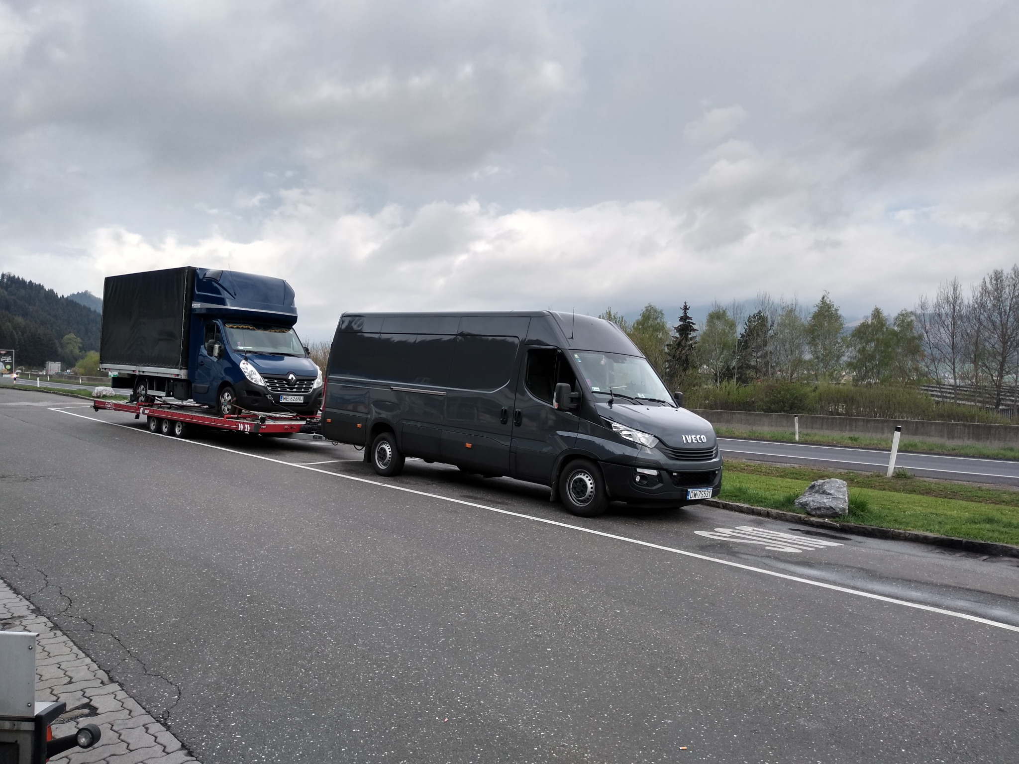 Ciemnoszara laweta Iveco z niebieskim samochodem dostawczym na czerwonej platformie, zaparkowana na poboczu drogi w pochmurny dzień.
