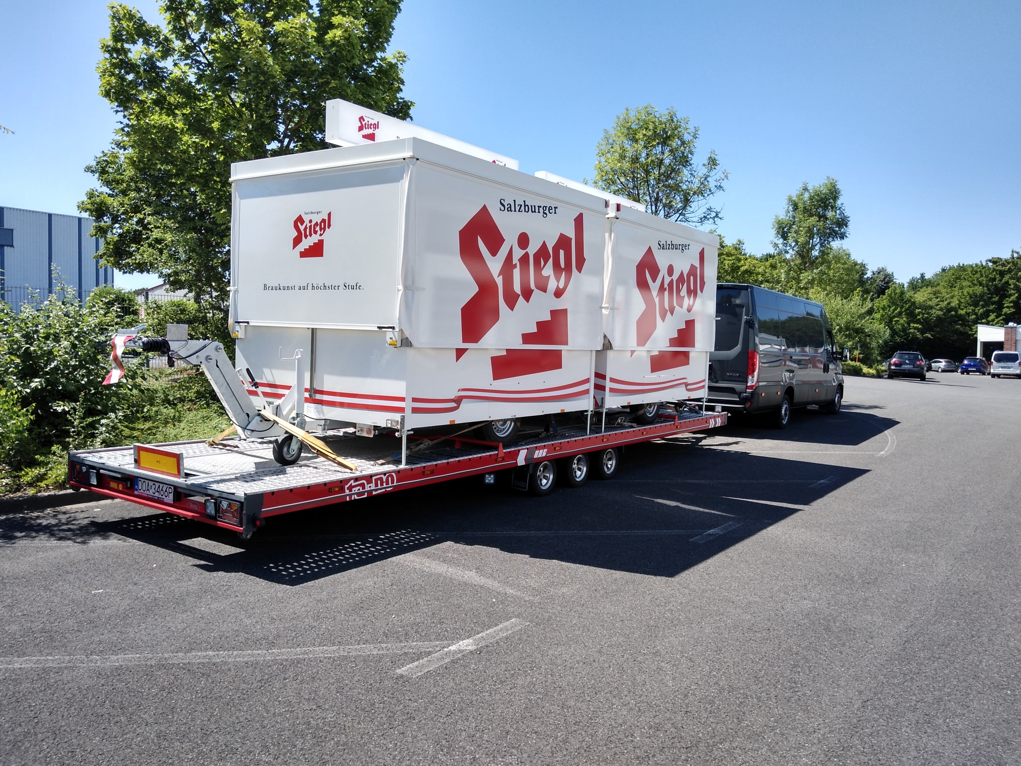 Czarny van ciągnie czerwoną lawetę z białą zabudową reklamową Stiegl na parkingu w słoneczny dzień.