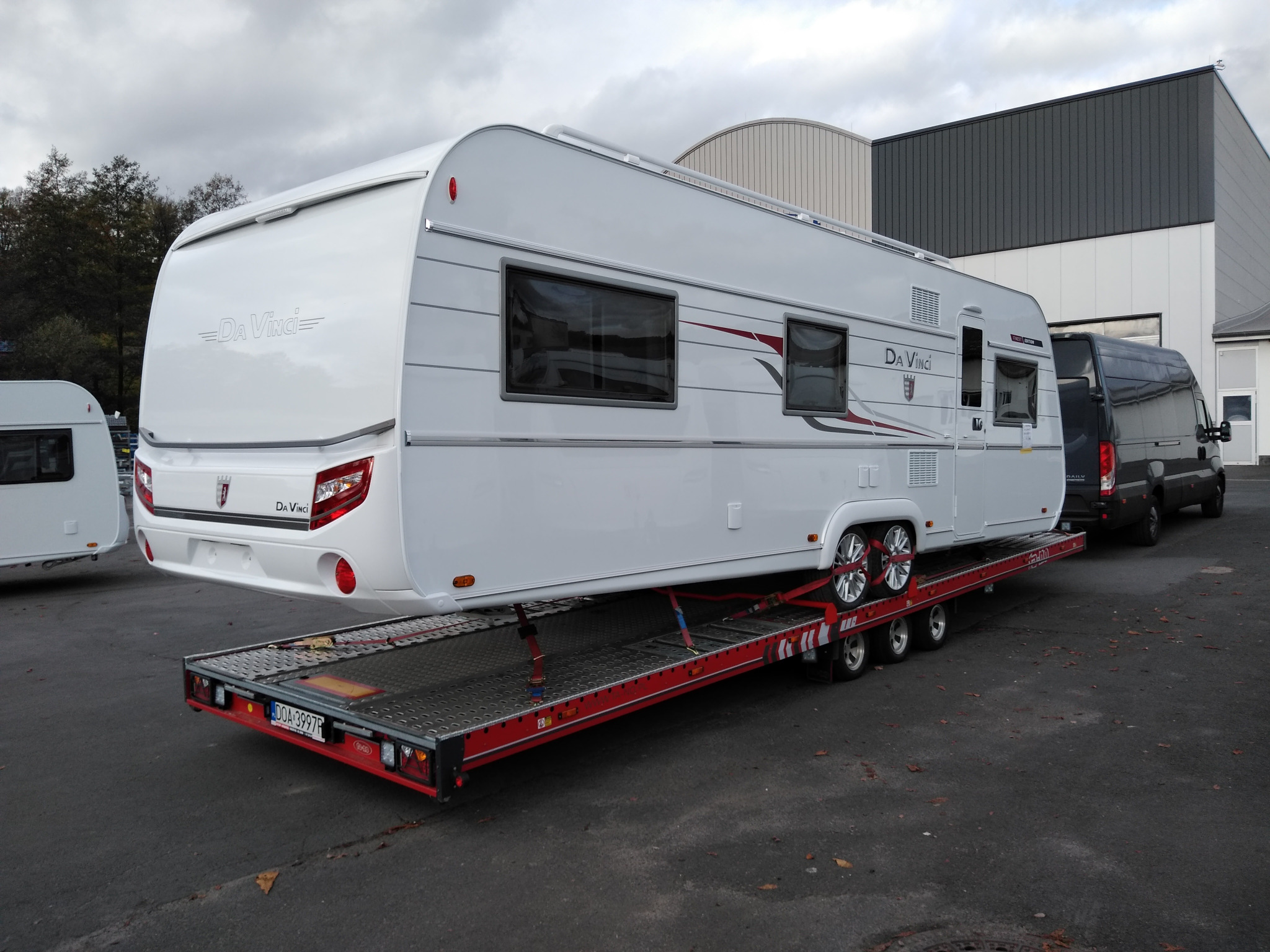 Biała przyczepa kempingowa marki Da Vinci umieszczona na czerwonej platformie lawety z widocznymi pasami zabezpieczającymi, zaparkowana na zewnątrz budynku przemysłowego.