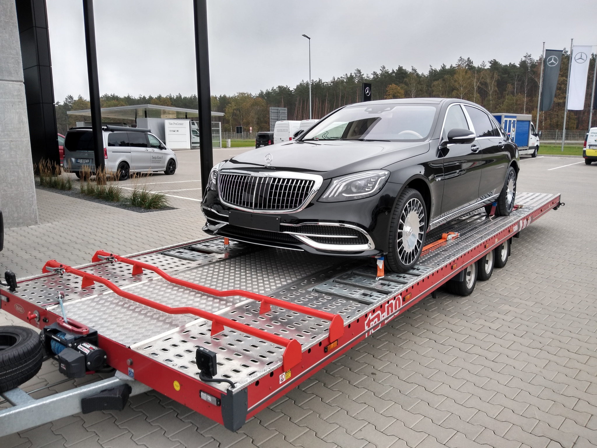 Czarny luksusowy samochód marki Mercedes-Benz Maybach zabezpieczony pasami na czerwonej platformie lawety na parkingu przed salonem samochodowym.