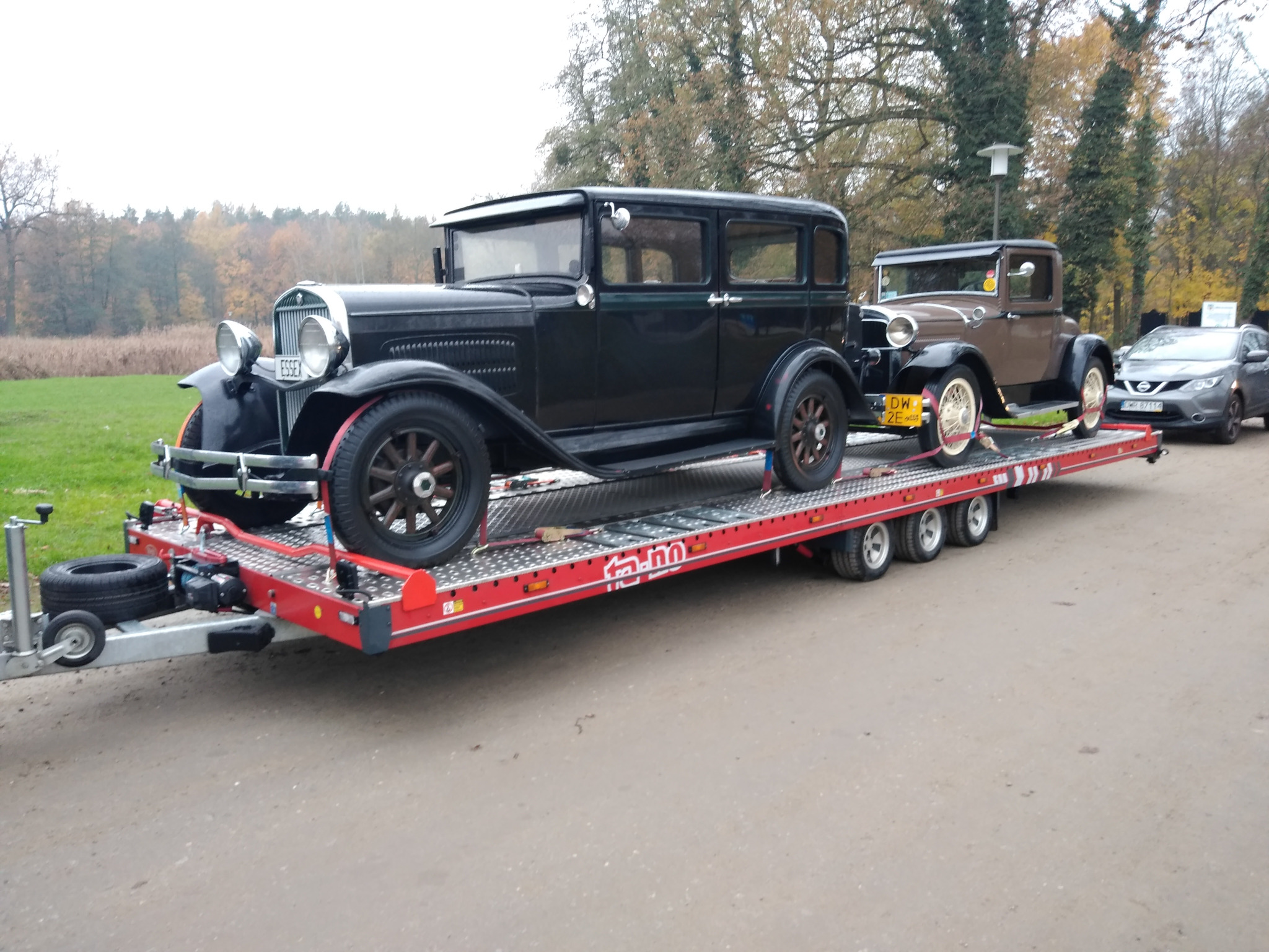 Dwa zabytkowe samochody, czarny Essex i brązowy Ford, zabezpieczone pasami na czerwonej lawecie z wieloma osiami, transportowane po asfaltowej drodze w jesiennej scenerii.
