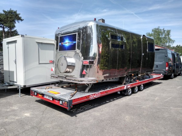 Błyszcząca, chromowana przyczepa kempingowa z ekranem reklamowym na tylnej ścianie, zabezpieczona pasami na czerwonej platformie lawety, obok zaparkowany bus.