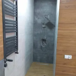 Wykończona łazienka z grzejnikiem drabinkowym w kolorze grafitowym, szarą strefą prysznicową z odpływem liniowym, płytkami imitującymi drewno na podłodze i ścianie, oraz białymi płytkami na ścianie...