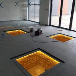 Podłoga z szarych płytek z trzema kwadratowymi otworami, w których widać cegły i oświetlenie LED, narzędzia i materiały budowlane w tle, niebieska taśma malarska na ścianie.