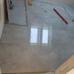 Adbud - Układanie szarych, matowych płytek podłogowych w łazience, widoczny poziomica, fragment toalety i okno odbijające się w powierzchni płytek.