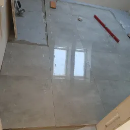 Układanie szarych, matowych płytek podłogowych w łazience, widoczny poziomica, fragment toalety i okno odbijające się w powierzchni płytek.