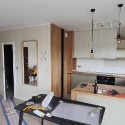 Adbud - Mężczyzna instaluje listwę przypodłogową przy wejściu do pokoju, widoczna nowa zabudowa kuchenna w tle, na pierwszym planie stół z narzędziami i oświetleniem LED.