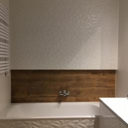 Adbud - Nowoczesna łazienka po remoncie: ściana z białych trójwymiarowych płytek, pas brązowych płytek imitujących drewno nad wanną, biała wanna z obudową z trójwymiarowych płytek, chromowana bateria.