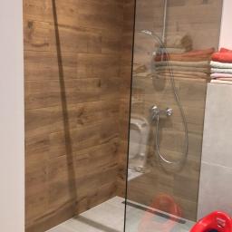 Adbud - Kabina prysznicowa ze szklaną ścianką, wyłożona płytkami imitującymi drewno, z widocznym odpływem liniowym i dziecięcą nakładką na sedes w rogu.