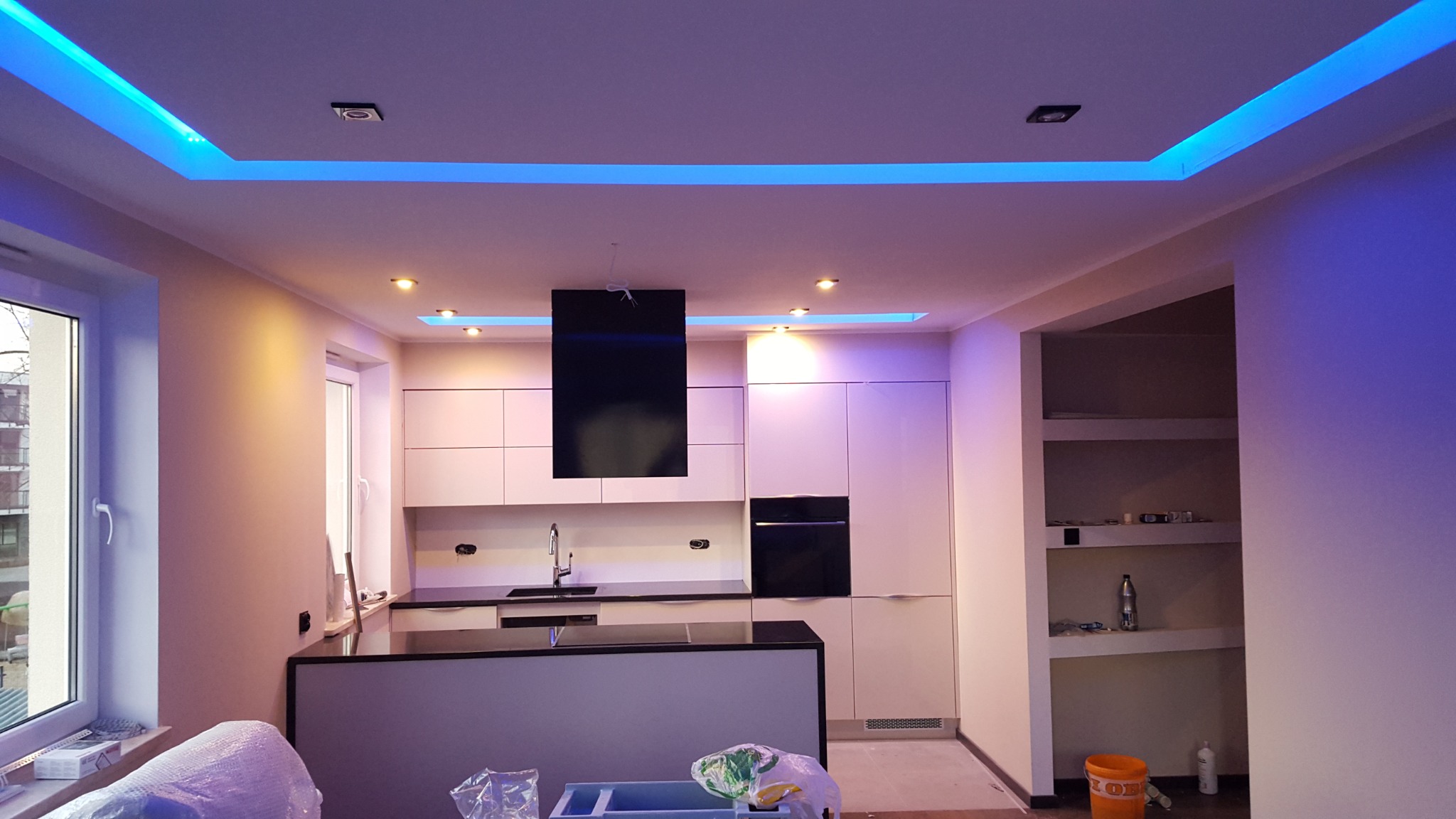 Nowoczesna kuchnia z białymi szafkami i czarnym blatem, podświetlonym sufitem na niebiesko, w trakcie wykańczania, widoczne narzędzia i materiały budowlane.