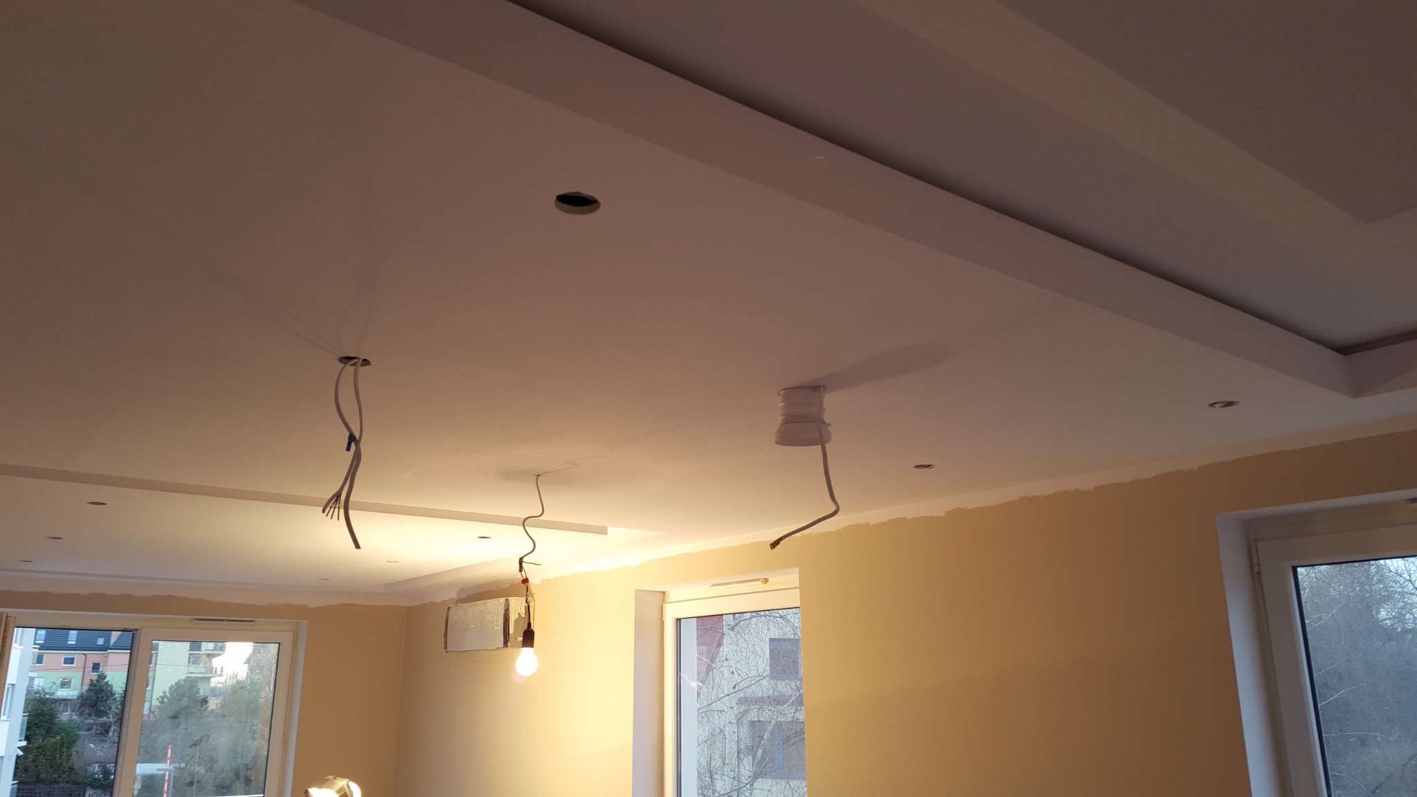 Niedokończony sufit podwieszany z wystającymi przewodami elektrycznymi i zamontowaną klimatyzacją, widok na okno i budynki za nim.