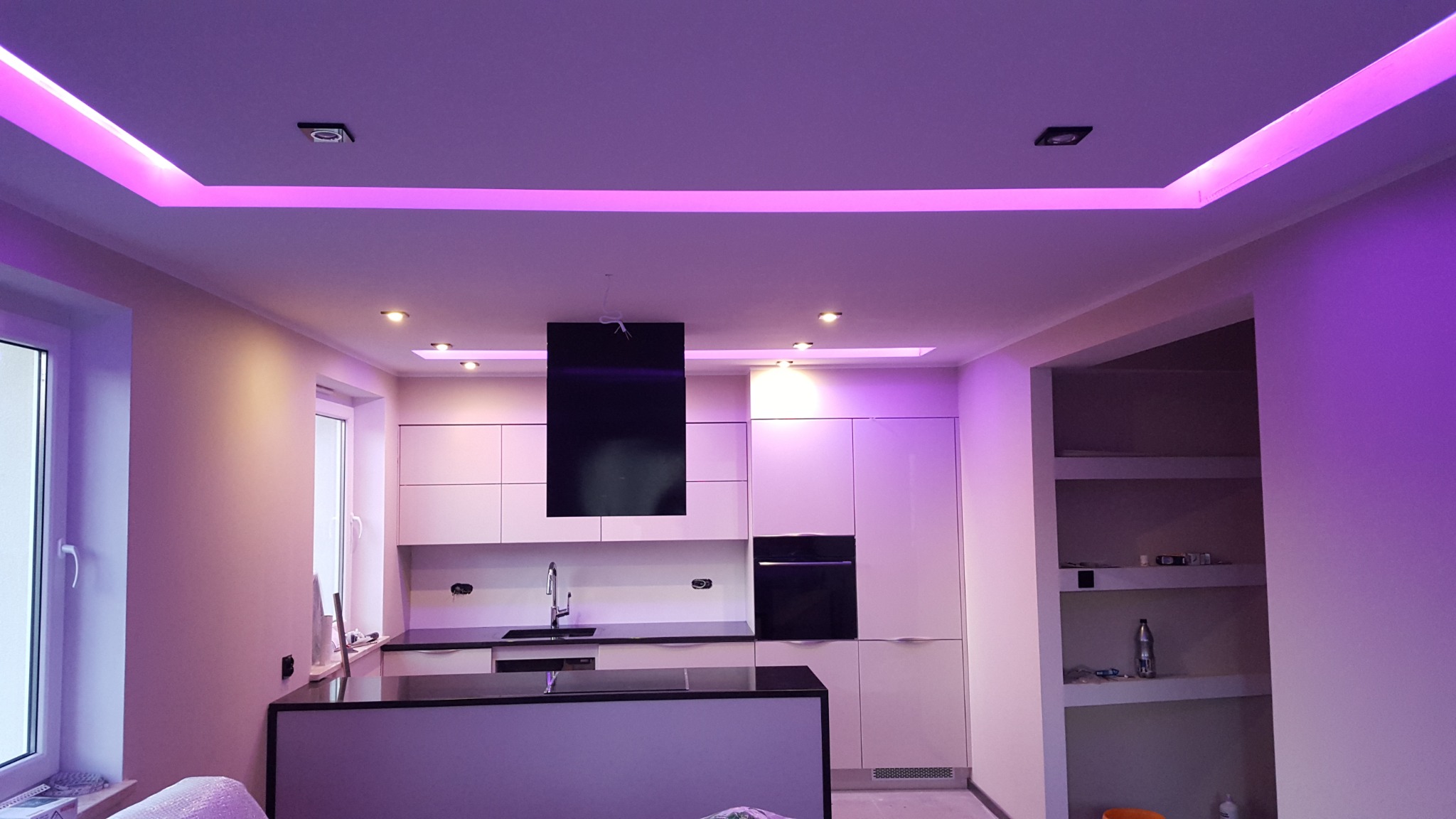 Nowoczesna kuchnia z białymi szafkami, czarnym blatem i okapem, oświetlona fioletowym światłem LED na suficie podwieszanym.