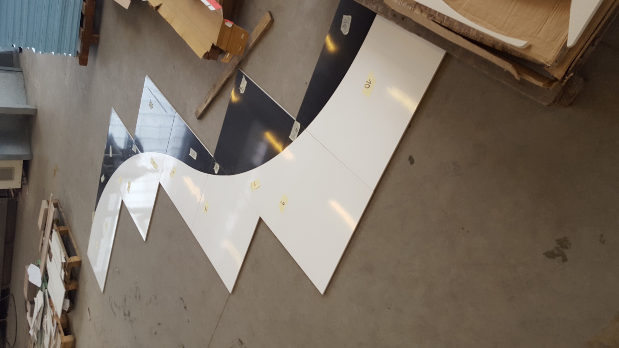 Elementy z białych i czarnych płyt, prawdopodobnie fragmenty blatu, ułożone na podłodze w falisty wzór, z numeracją na żółtych karteczkach, czekające na montaż.