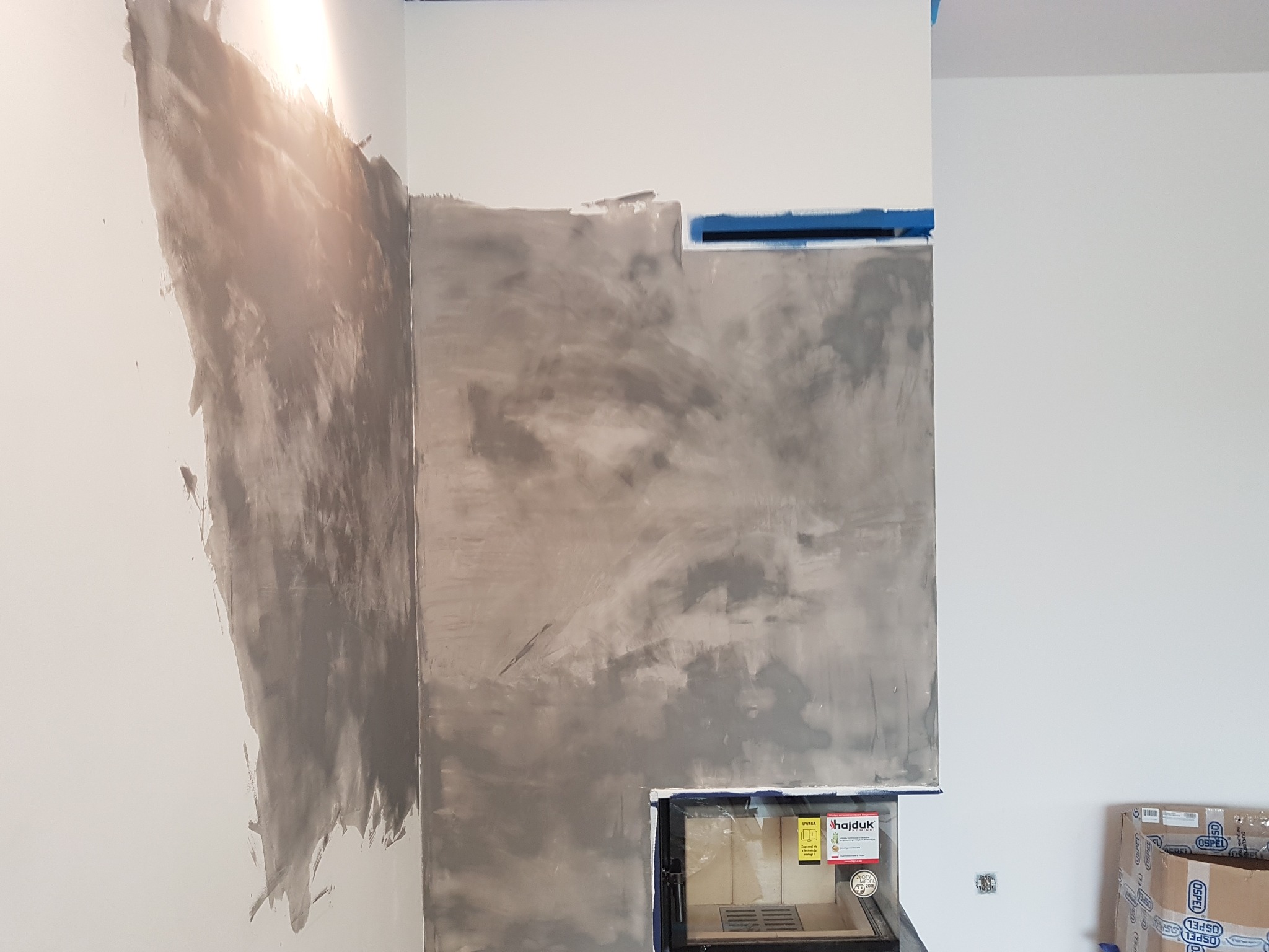 Ściana z nałożonym szarym tynkiem dekoracyjnym imitującym beton, zabezpieczona niebieską taśmą malarską, z zamontowanym wkładem kominkowym marki Hajduk z widoczną instrukcją obsługi na szybie.