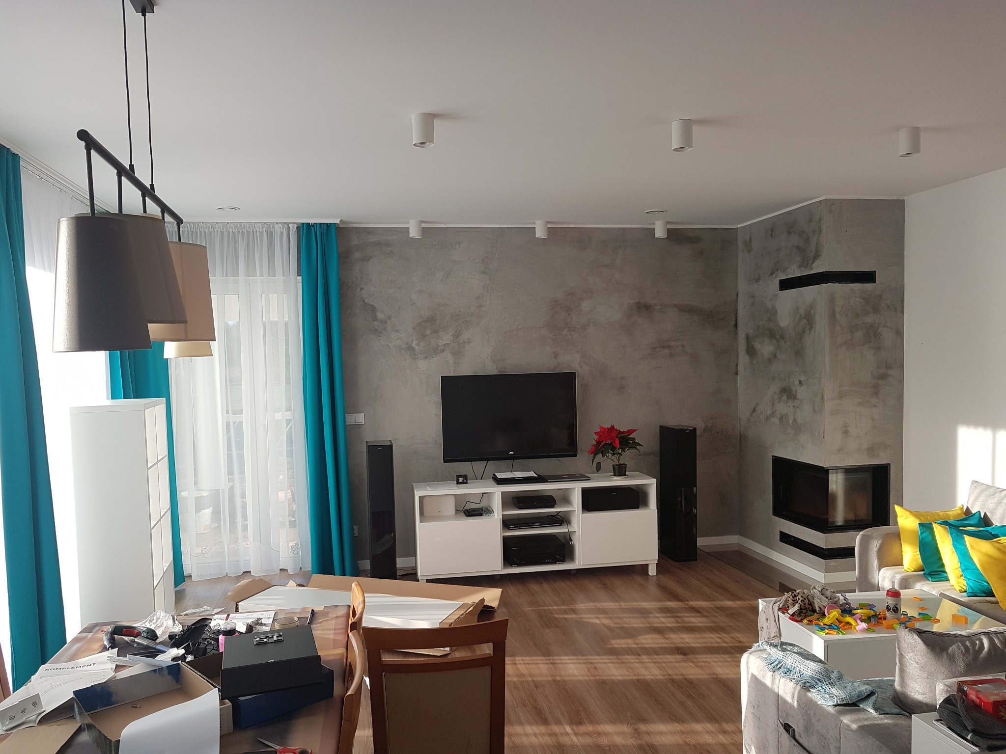Salon w trakcie aranżacji: szara ściana z efektem betonu, biała szafka RTV, telewizor, kominek narożny, turkusowe zasłony i rozpakowane elementy mebli na stole.