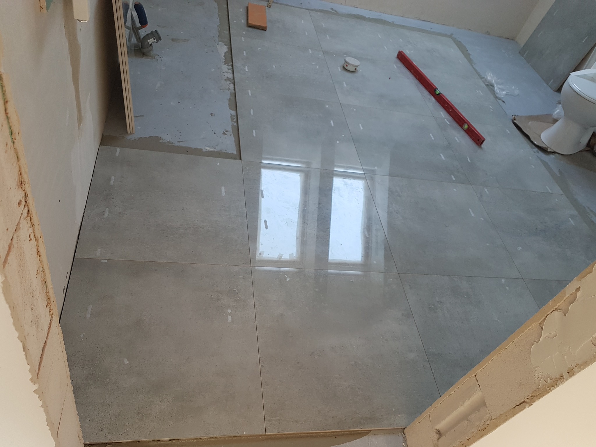 Układanie szarych, matowych płytek podłogowych w łazience, widoczny poziomica, fragment toalety i okno odbijające się w powierzchni płytek.