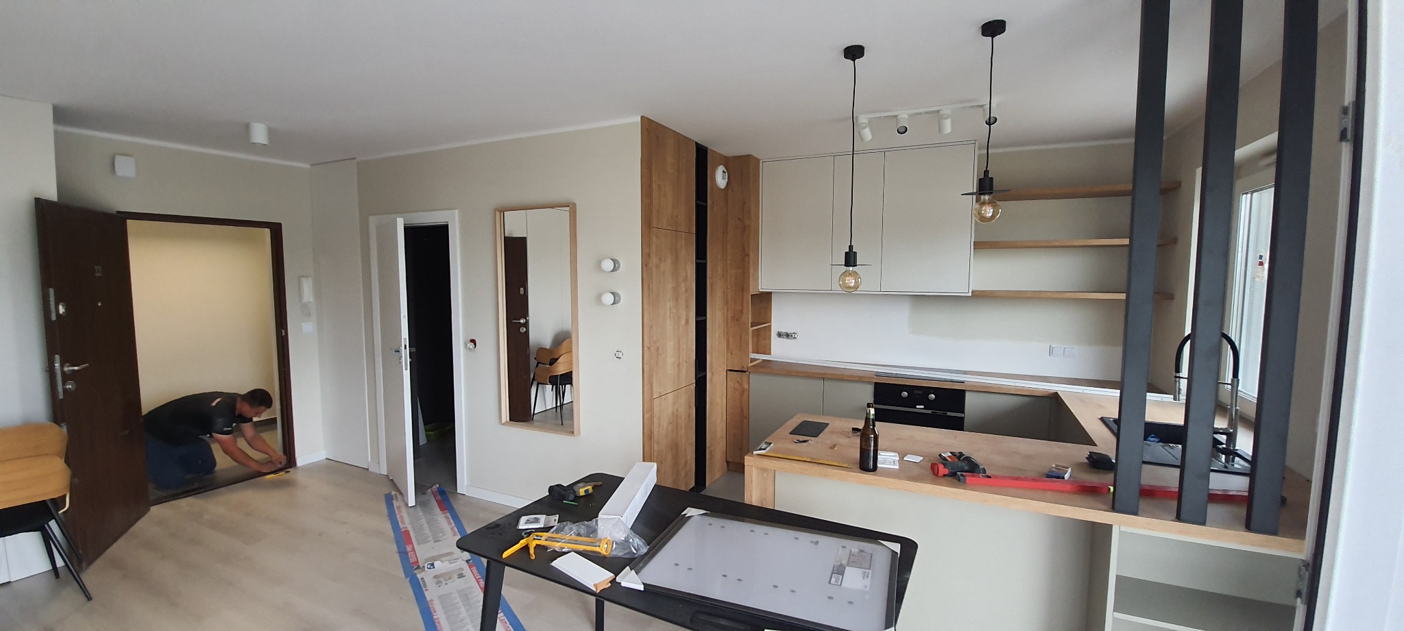 Mężczyzna instaluje listwę przypodłogową przy wejściu do pokoju, widoczna nowa zabudowa kuchenna w tle, na pierwszym planie stół z narzędziami i oświetleniem LED.