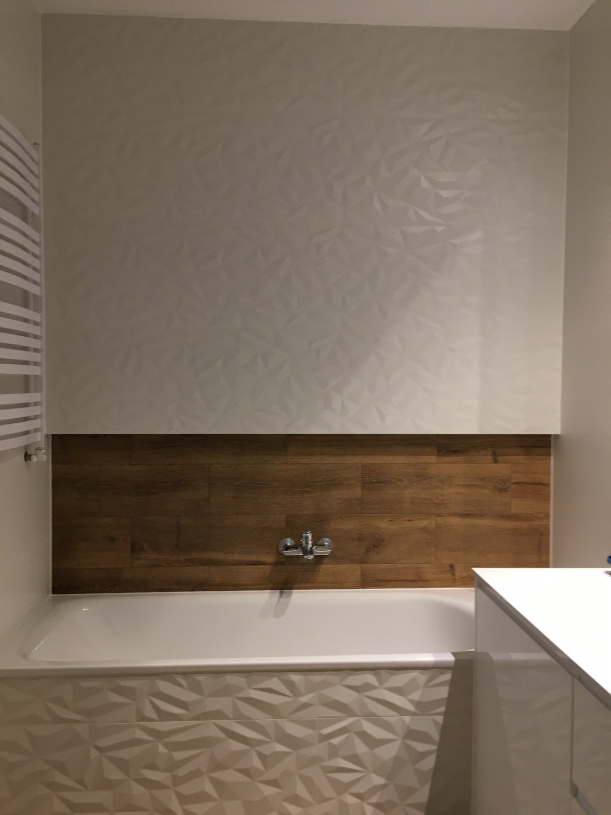 Nowoczesna łazienka po remoncie: ściana z białych trójwymiarowych płytek, pas brązowych płytek imitujących drewno nad wanną, biała wanna z obudową z trójwymiarowych płytek, chromowana bateria.