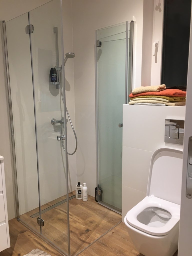 Nowoczesna łazienka z kabiną prysznicową ze szkła hartowanego, drewnopodobną podłogą, wiszącą toaletą i stosem ręczników w ciepłych kolorach obok okna.