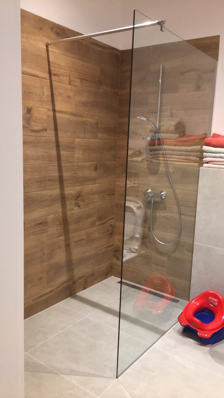 Kabina prysznicowa ze szklaną ścianką, wyłożona płytkami imitującymi drewno, z widocznym odpływem liniowym i dziecięcą nakładką na sedes w rogu.