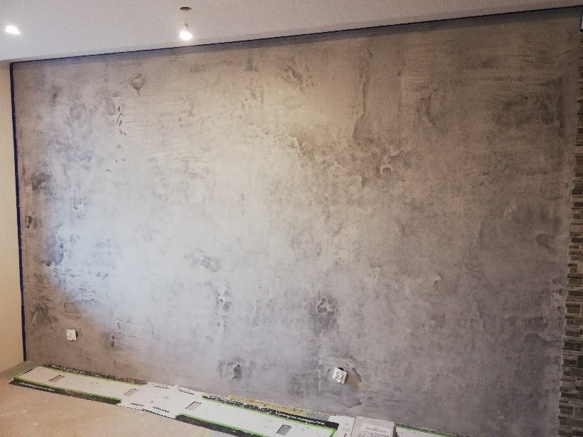 Ściana w pokoju pokryta dekoracyjnym tynkiem w odcieniach szarości i beżu, z widocznymi elementami instalacji elektrycznej i fragmentem ściany wykończonej płytkami imitującymi cegłę po prawej...