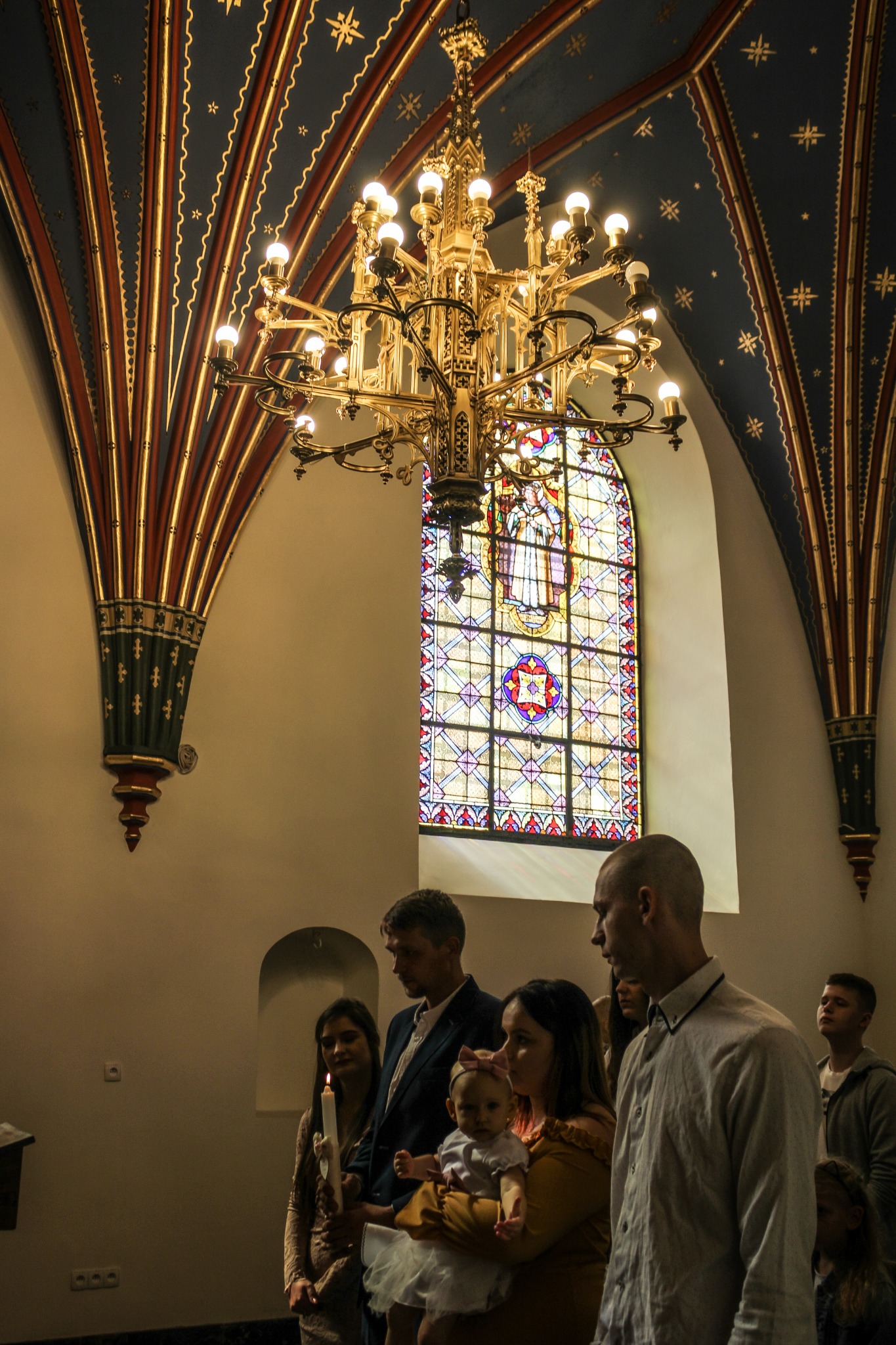 Rodzina z małym dzieckiem w kościele, pod bogato zdobionym żyrandolem i witrażem, podczas uroczystości religijnej.