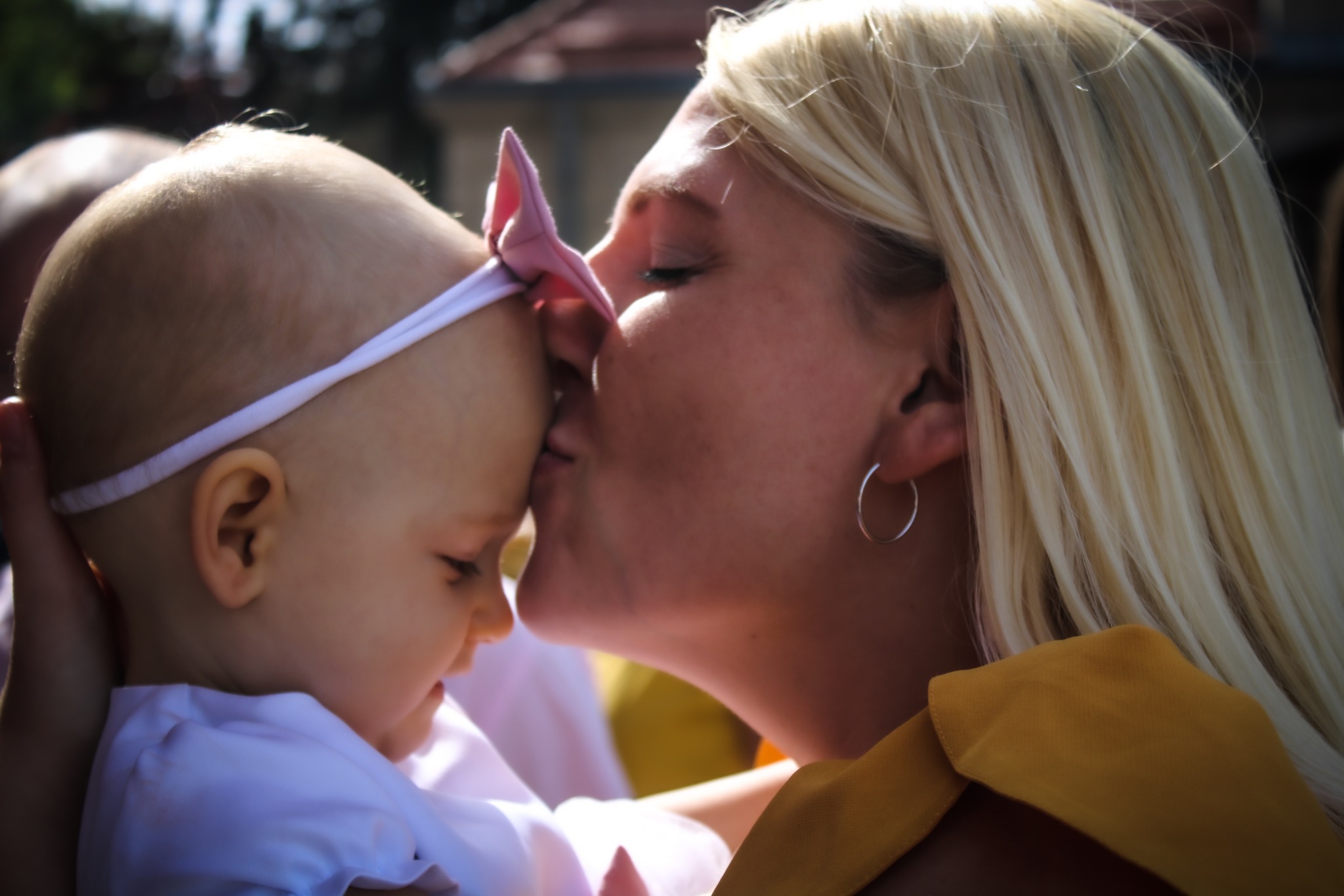 Mama całuje w czoło niemowlę z różową kokardką na głowie, widoczne srebrne kolczyki mamy i musztardowa bluzka.