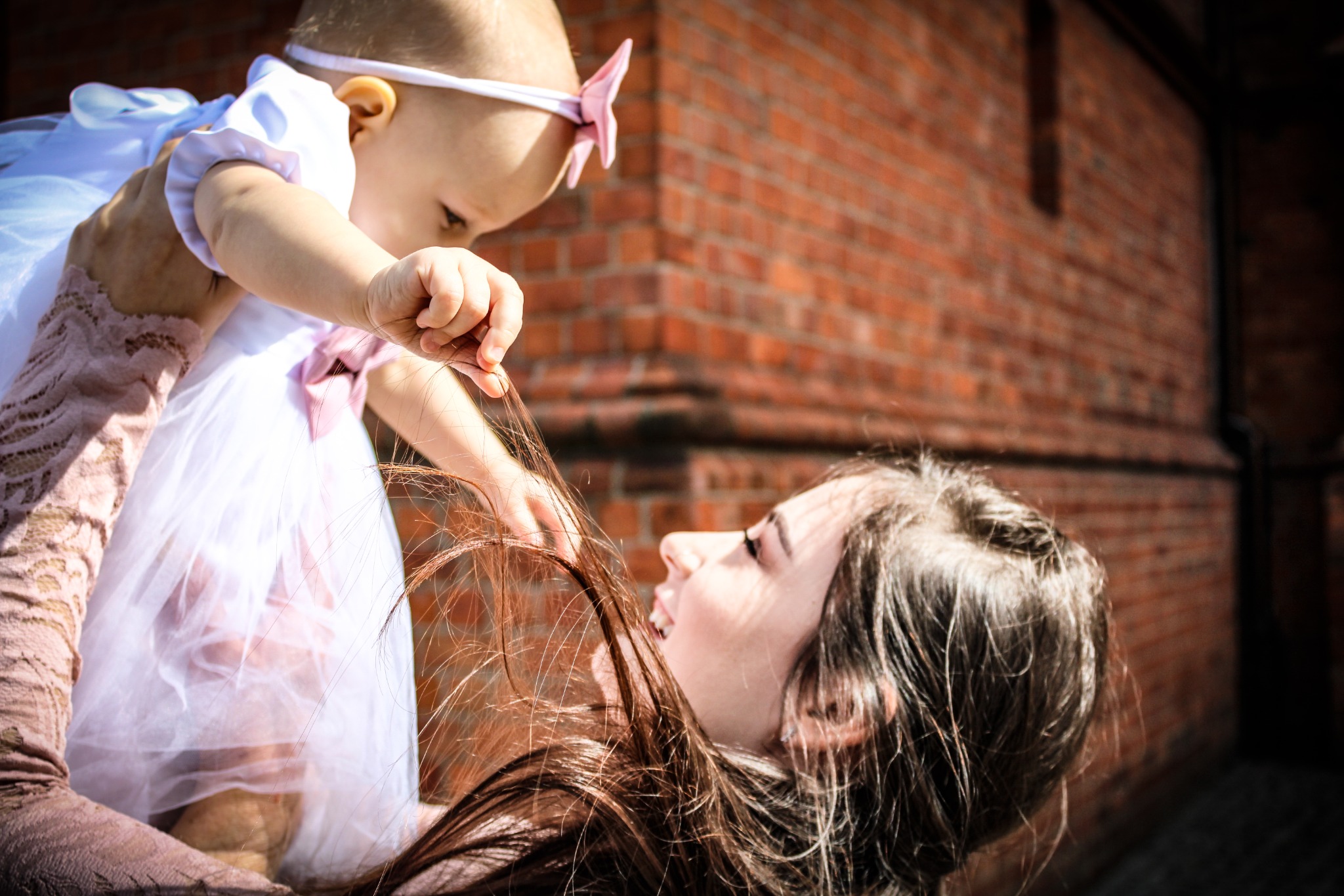 Uśmiechnięta mama trzymająca w górze niemowlę w białej sukience z różową kokardką na głowie, dziecko bawi się włosami matki na tle ceglanej ściany.