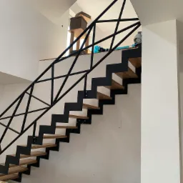 Nowoczesne schody metalowe z drewnianymi stopniami i geometryczną balustradą, widoczne mocowania do ściany, w tle drapak dla kota i lampa wisząca.