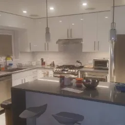 Biała kuchnia z połyskiem, wyspa kuchenna z szarym blatem, nowoczesne oświetlenie punktowe i wiszące lampy, stalowa lodówka i sprzęt AGD, widok na okno z roślinami.