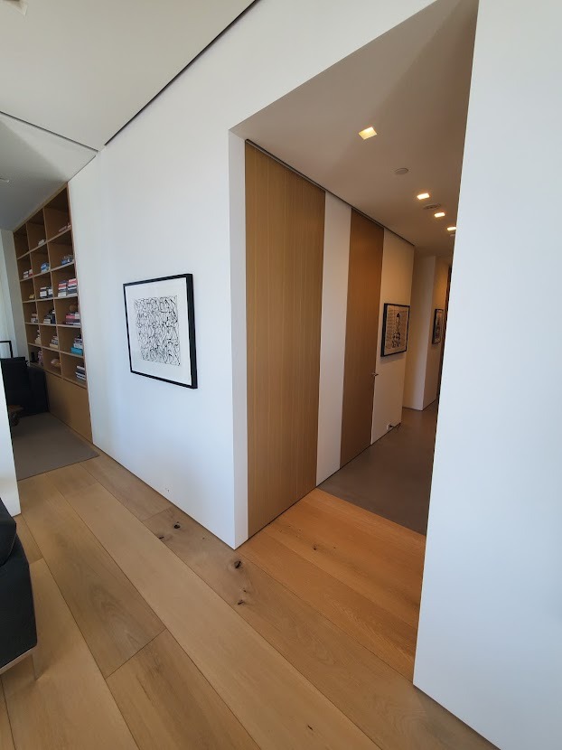 Nowoczesne wnętrze z drewnianą zabudową meblową na wymiar, obejmującą regał z książkami i ukryte drzwi w panelach ściennych, z minimalistycznym obrazem na ścianie.