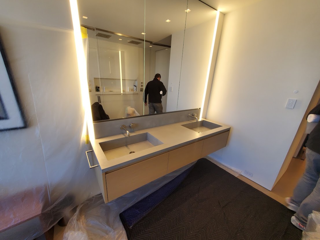 Nowoczesna łazienka z podwójną umywalką w betonowym blacie, podświetlane lustro i widoczne prace wykończeniowe.