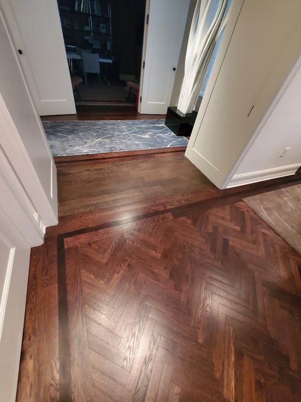 Ułożona w jodełkę, ciemna drewniana podłoga z bordiurą, widoczna z perspektywy wejścia do pokoju z dywanem w odcieniach szarości i bieli, oraz dekoracyjną rzeźbą przy ścianie.