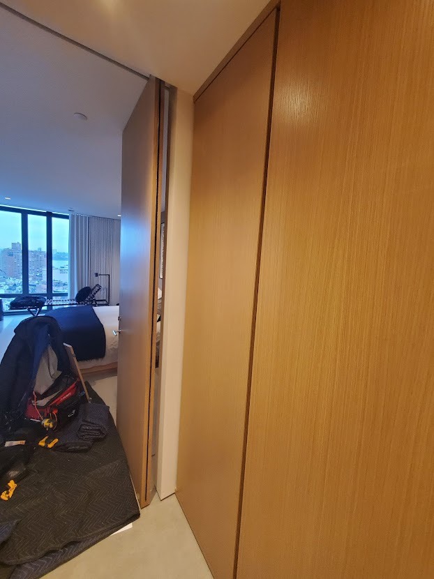 Montaż przesuwnych drzwi szafy wnękowej z drewnianym fornirem, widoczne narzędzia i materiały zabezpieczające podłogę, w tle widok na miasto przez okno.