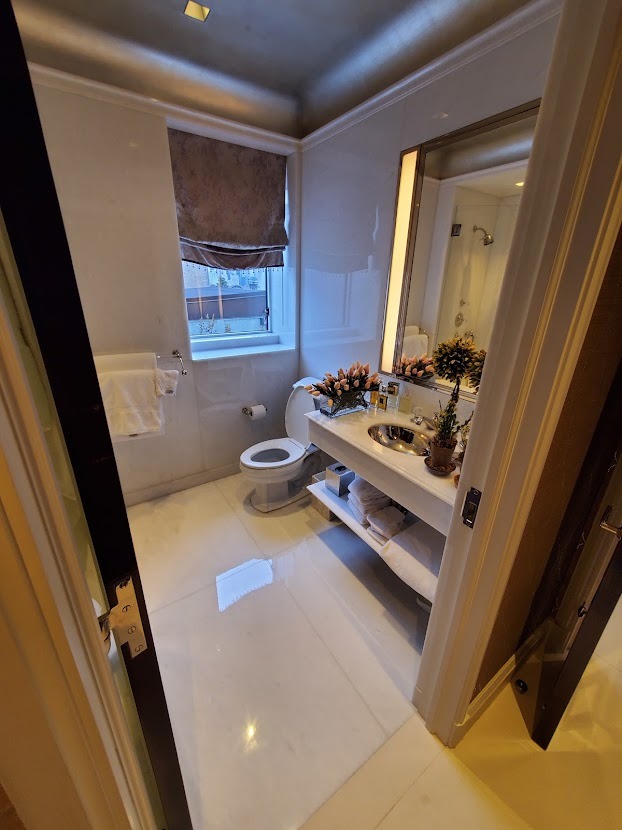 Elegancka łazienka z marmurowymi ścianami, oknem z zasłoną rzymską i toaletką z umywalką, kwiatami i kosmetykami, widok zza uchylonych drzwi.